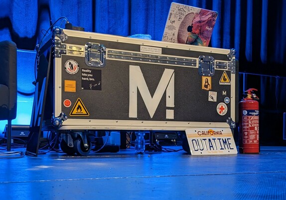 Eine Transportkiste mit dem Methodisch Inkorrekt Symbol steht auf einer Bühne. Davor steht ein Nummernschild mit der Aufschrift Outatime. Auf der Kiste steht ein Laptop und daneben ein Feuerlöscher. 